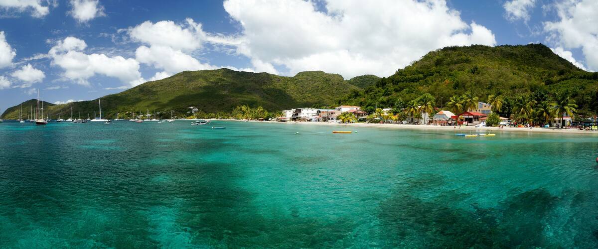 A tropical beach with clear turquoise waters, a small town with buildings and palm trees, and a mountain in the background.