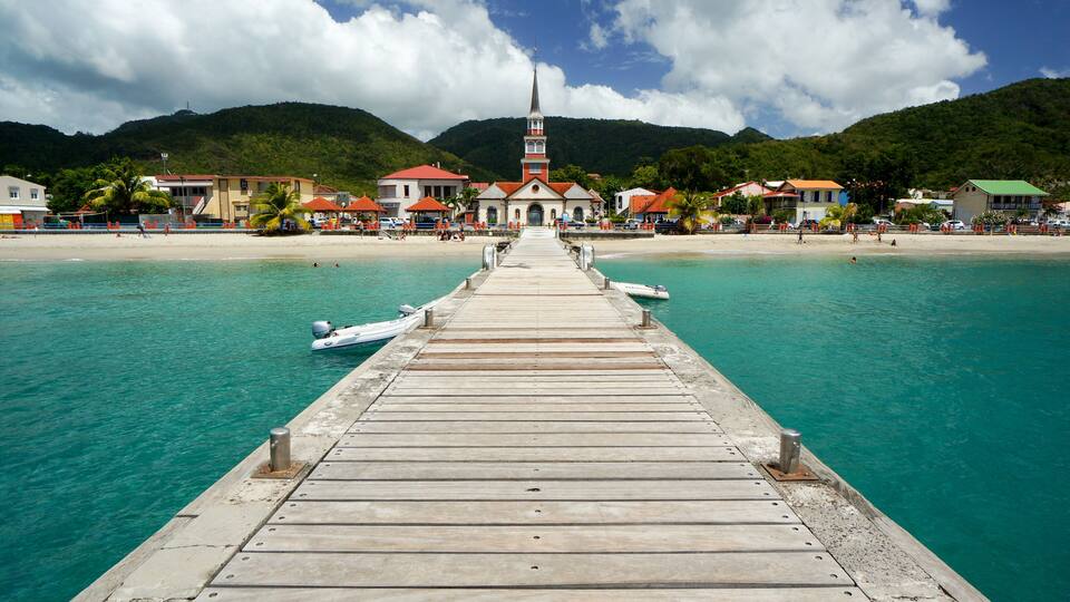 A wooden pier leading to a church with a tall steeple, a beach, and mountains in the background.
