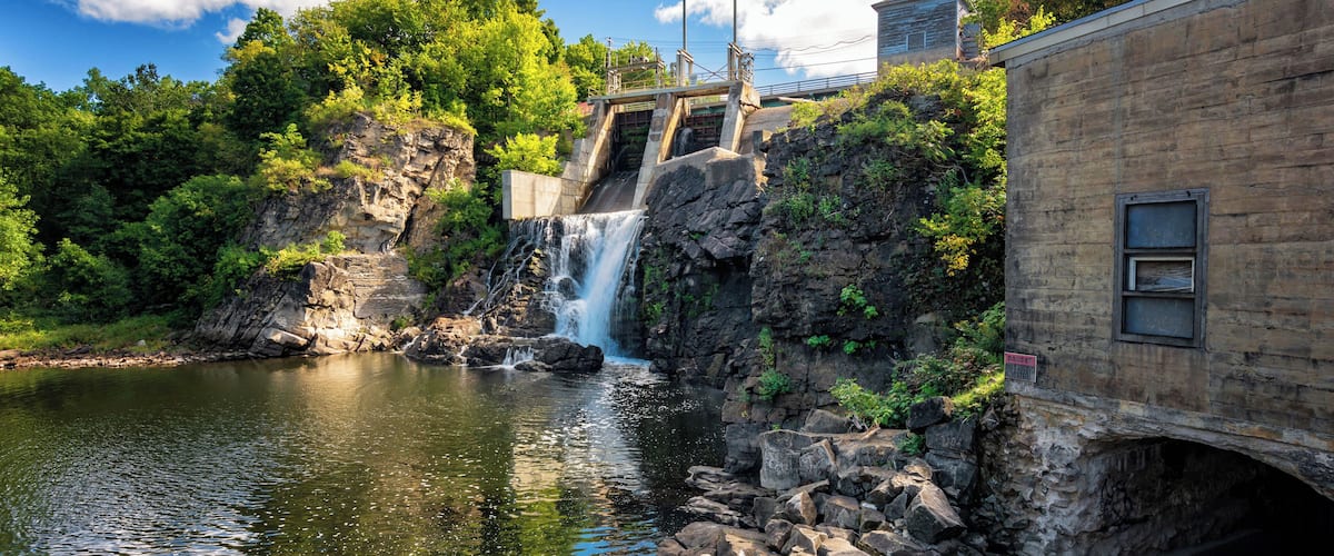 A waterfall cascading over rocks into a river, surrounded by lush greenery and a bridge.