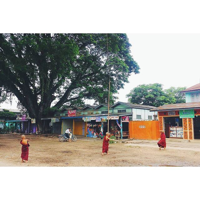 Una calle con un árbol grande, tiendas y dos personas vestidas con trajes tradicionales.