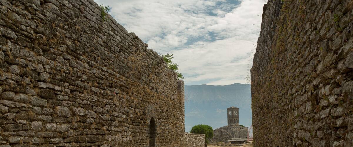 A stone pathway flanked by ancient walls leading to a tower with a clock.