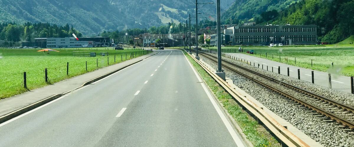 A road with a railway track, surrounded by mountains and greenery.