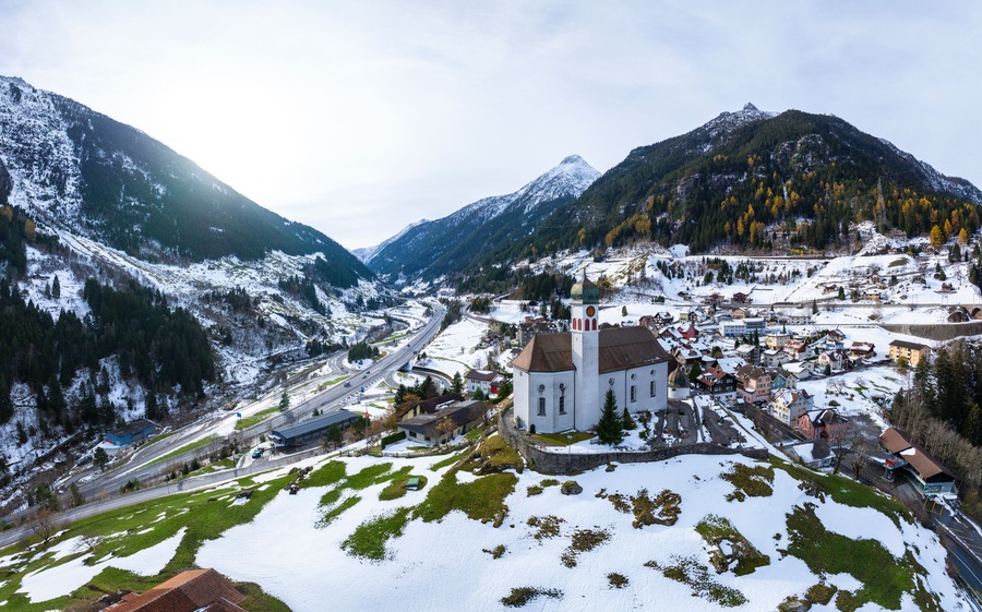Un pueblo cubierto de nieve en la montaña, con una iglesia destacada, carreteras sinuosas y un valle.