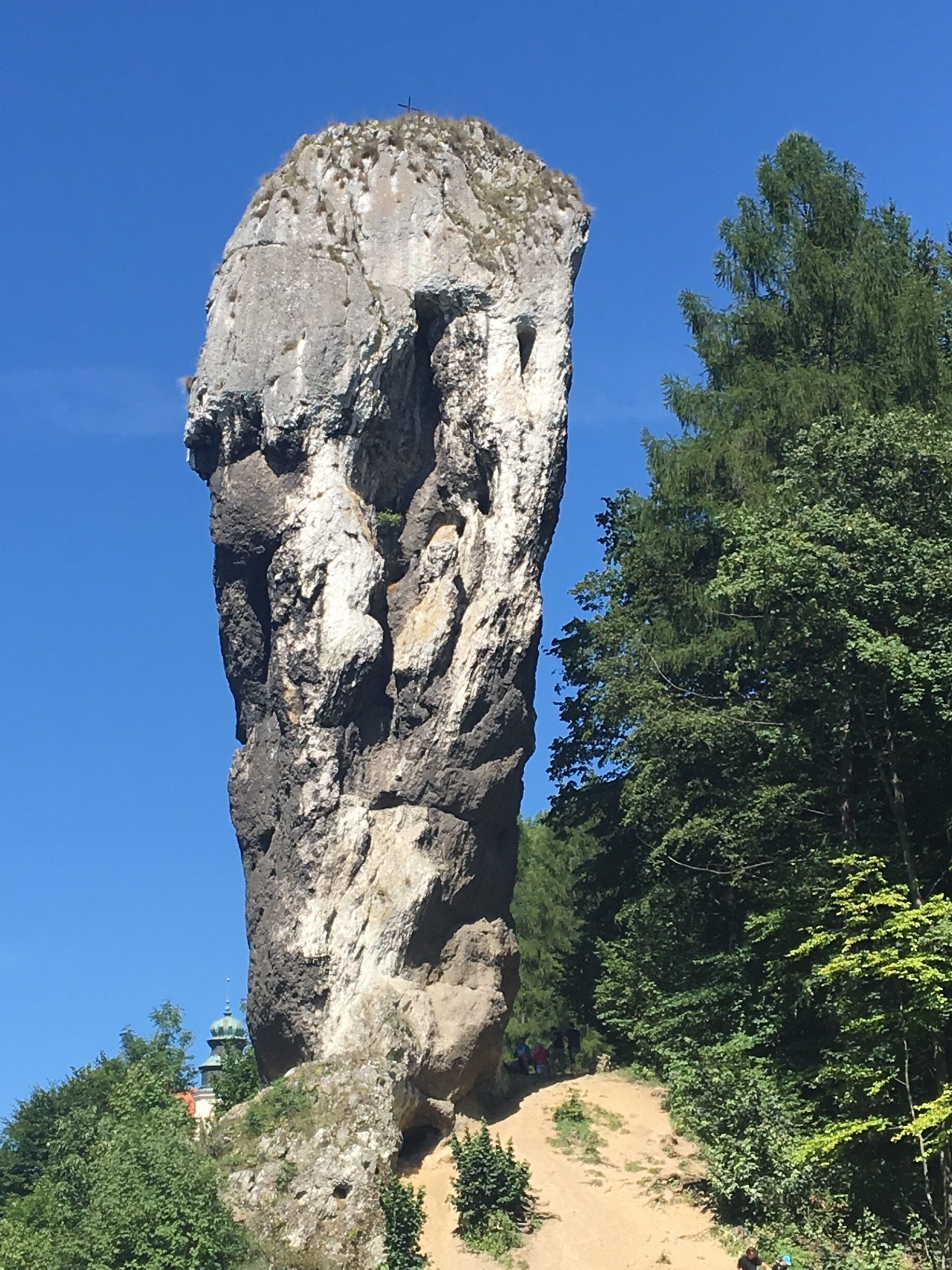 A tall rock formation with a cross on top, surrounded by trees and a clear blue sky.