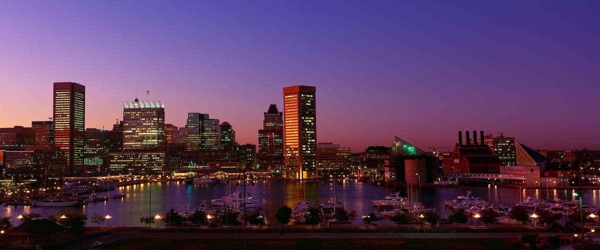 A city skyline at dusk with illuminated buildings and a marina with boats.