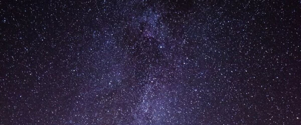 A starry night sky with a visible Milky Way, a road leading into the forest, and a silhouette of a person.