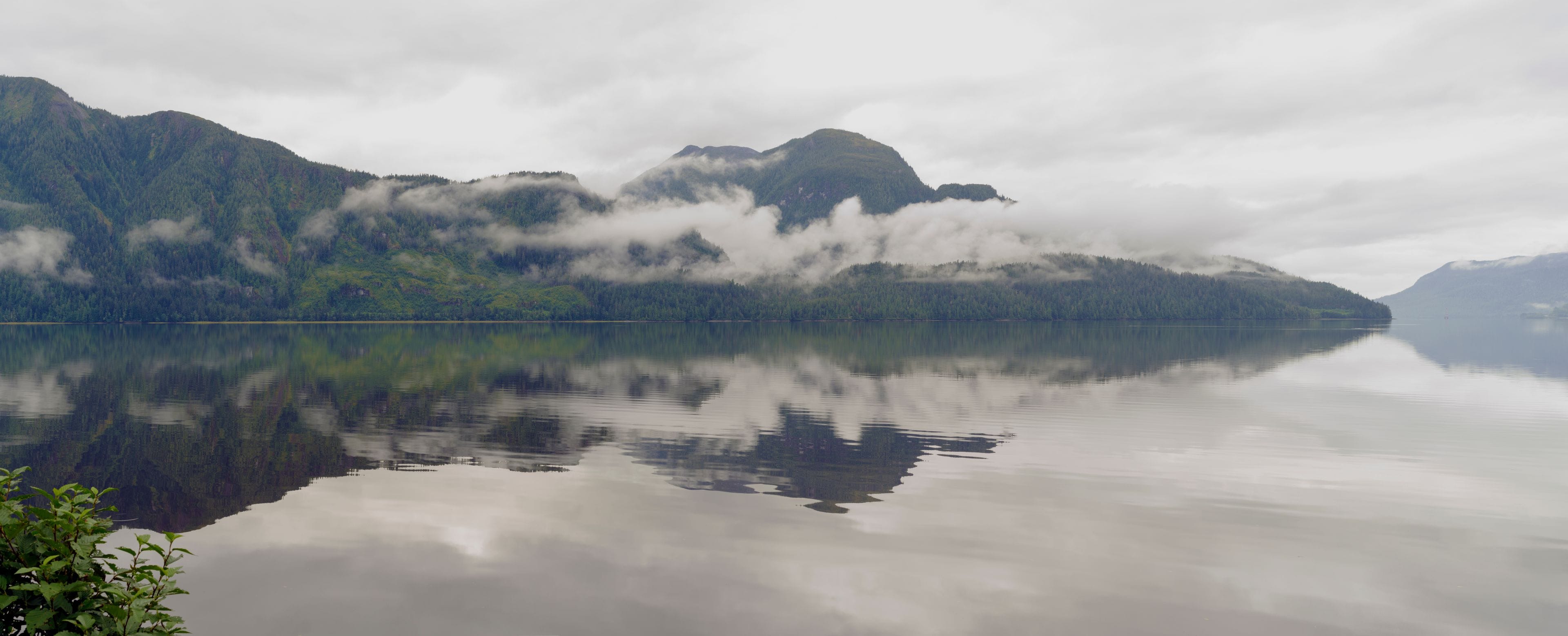 Un lago tranquilo con montañas al fondo, parcialmente cubiertas de nubes.
