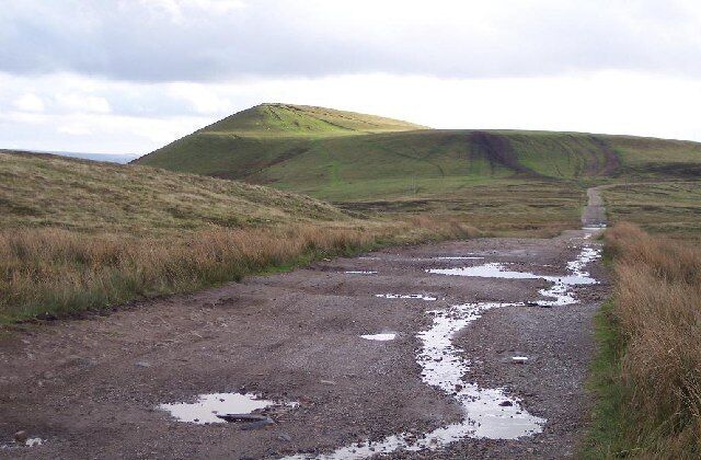 Une route de campagne parsemée de flaques d’eau, bordée de collines verdoyantes et surplombée par un ciel nuageux.
