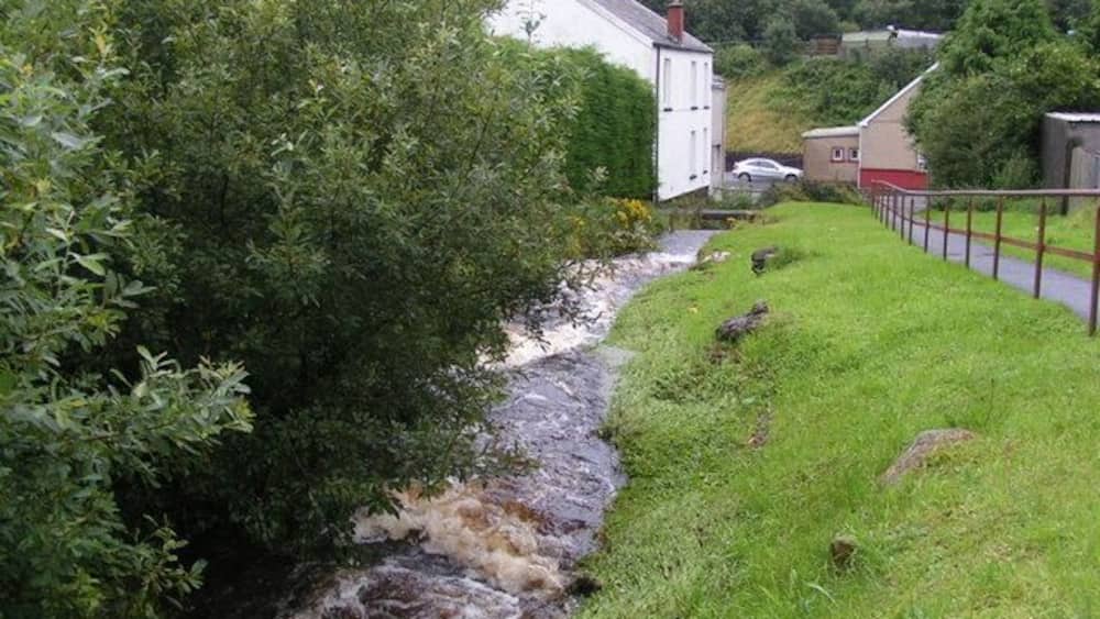 Une rivière traverse un paysage verdoyant, avec un bâtiment blanc et une clôture en arrière-plan.