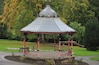 A gazebo with a glass roof and ornate metalwork, surrounded by a red and black fence, set in a park with trees and a bench.