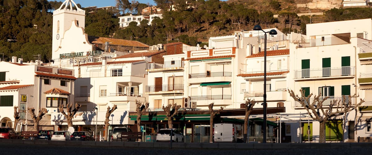 A coastal town with white buildings, a church tower, and parked cars along the street.