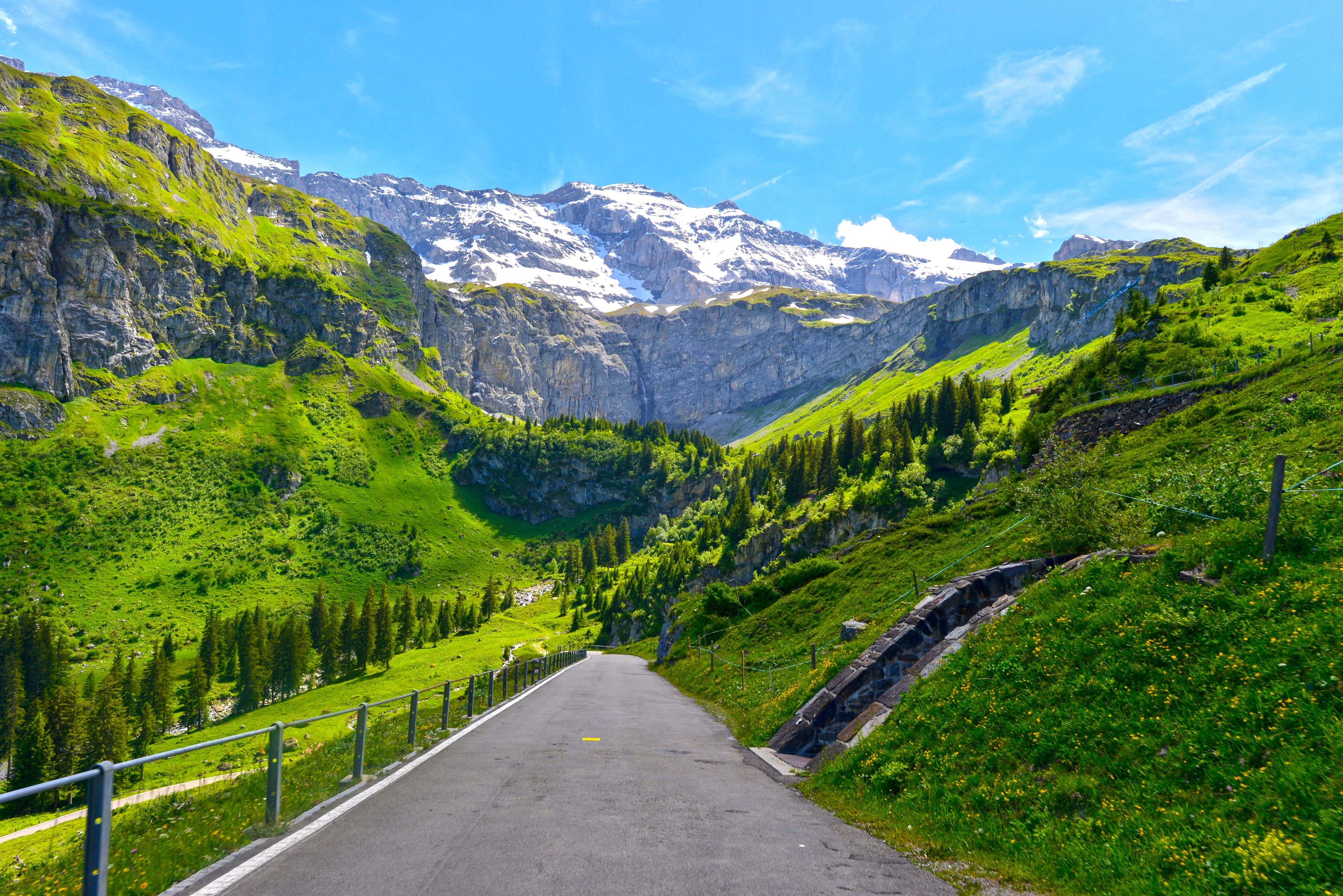 Une route de montagne bordée de collines verdoyantes et de sommets enneigés, le tout sous un ciel bleu dégagé.