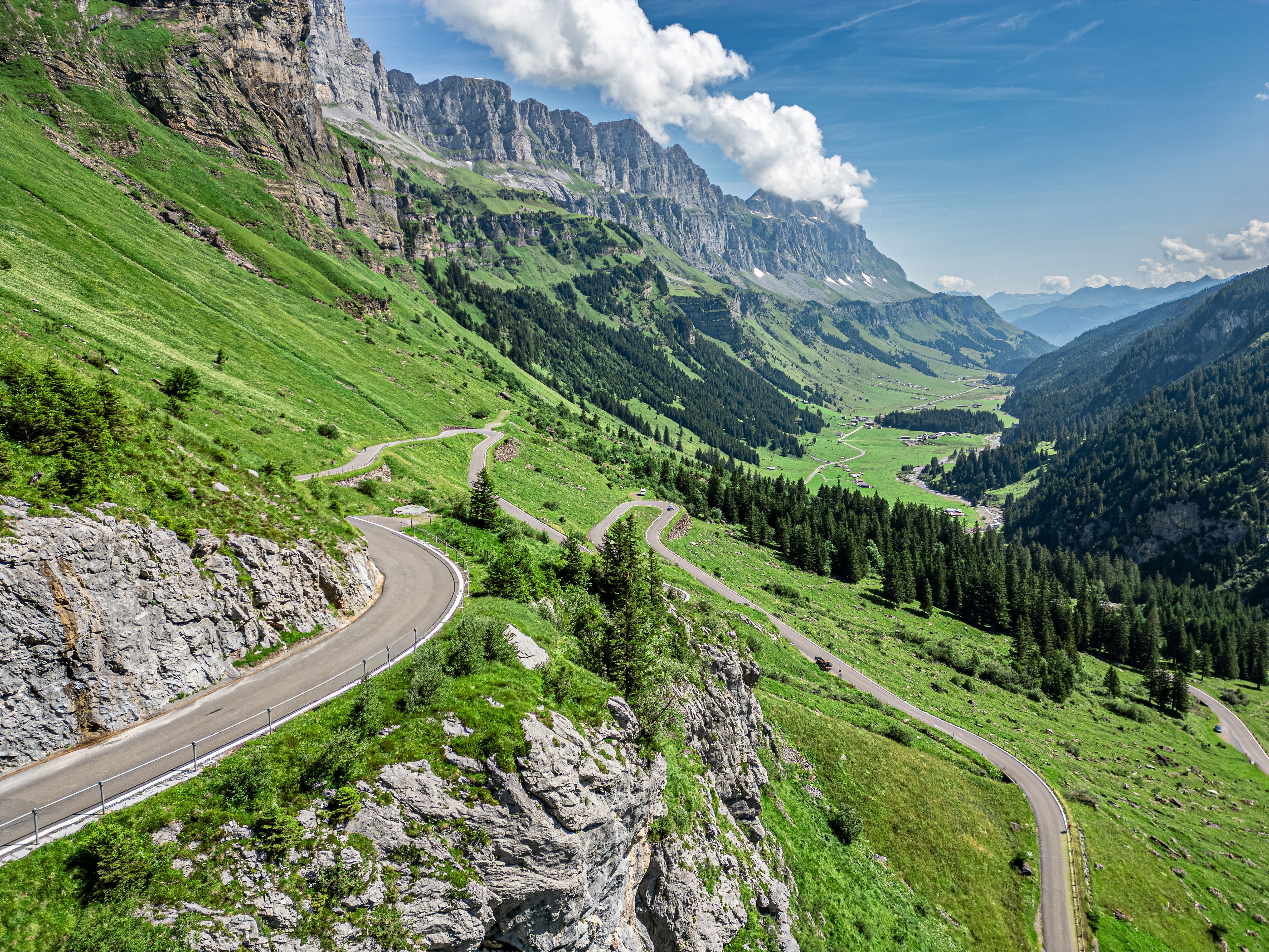 Une route de montagne sinueuse traverse une vallée verdoyante, bordée de falaises rocheuses et dominée par les sommets lointains des montagnes, le tout sous un ciel dégagé.