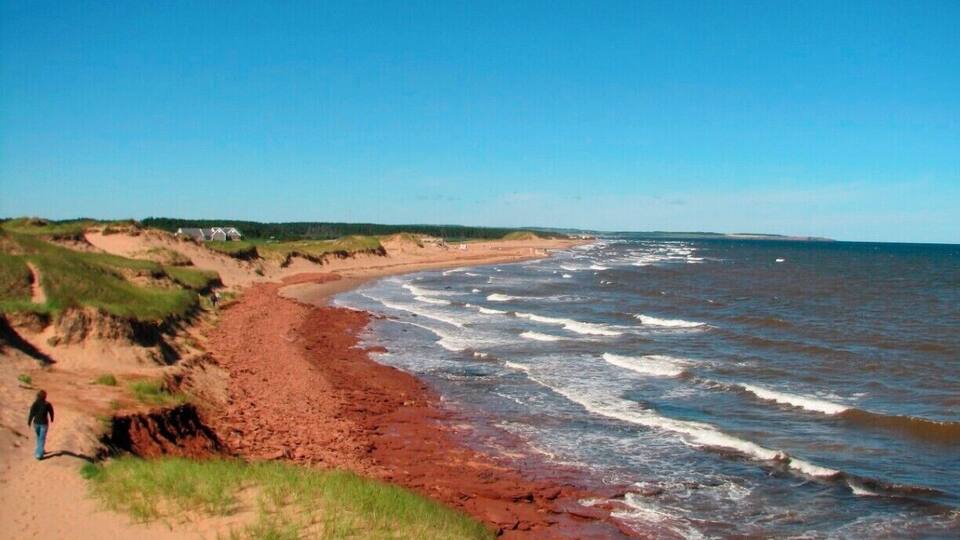 A coastal landscape with a red sandy beach, waves crashing onto the shore, and a person walking along the beach.
