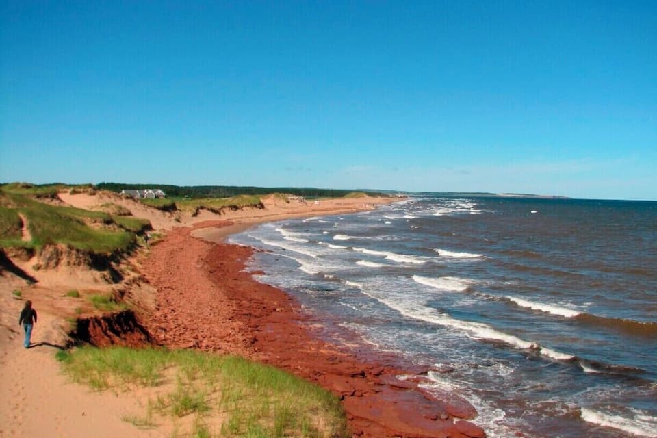 Et kystlandskab med en røget sandstrand, bølger der slår ind mod kysten, og en person der går en tur langs stranden.