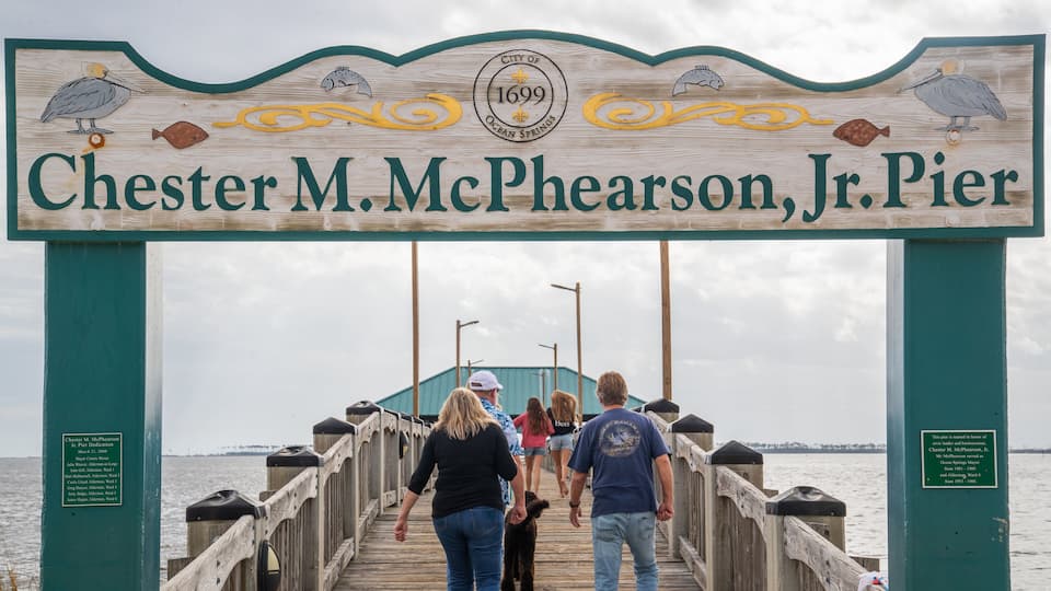 A pier named Chester M. McPheerson, Jr. Pier with a wooden sign featuring pelicans and fish, and a plaque commemorating its dedication.