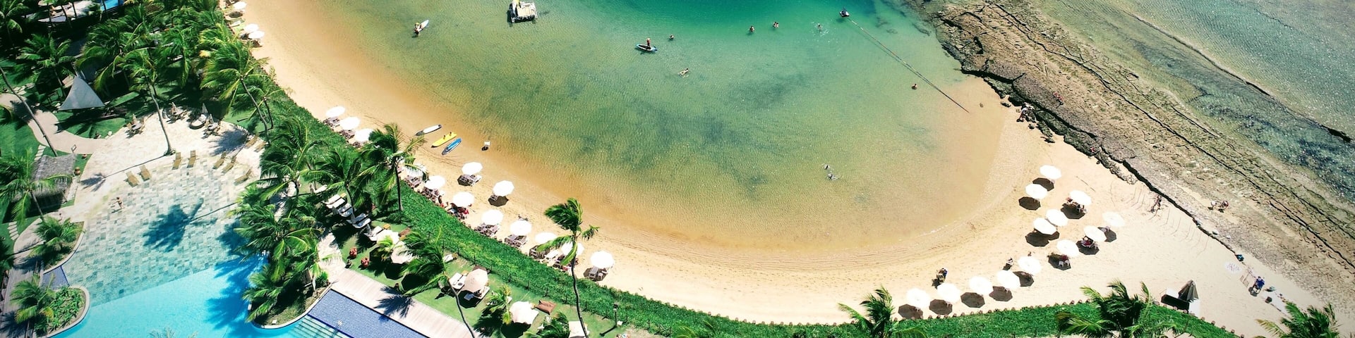 A coastal beach with clear turquoise waters, a sandy shoreline, and a resort area with umbrellas and palm trees.