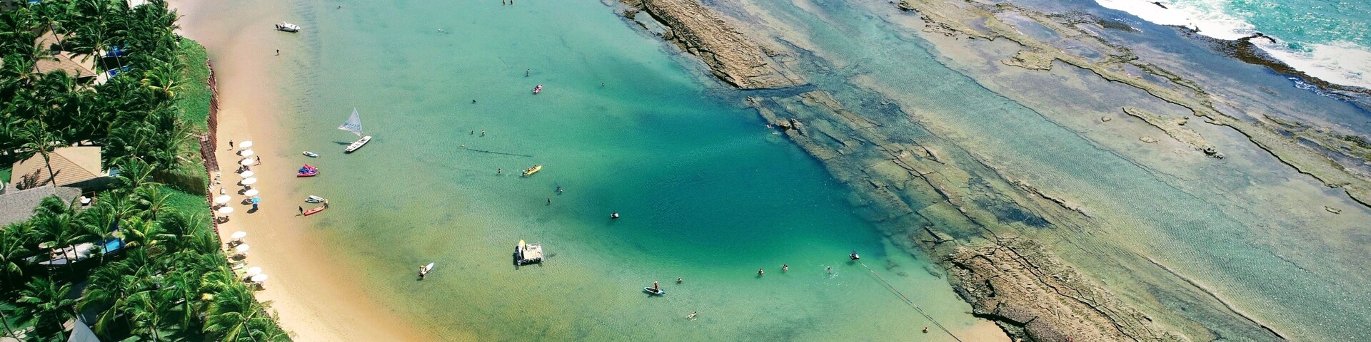 Uma praia costeira com águas turquesa cristalinas, areal arenoso e área de resort com guarda-sóis e palmeiras.