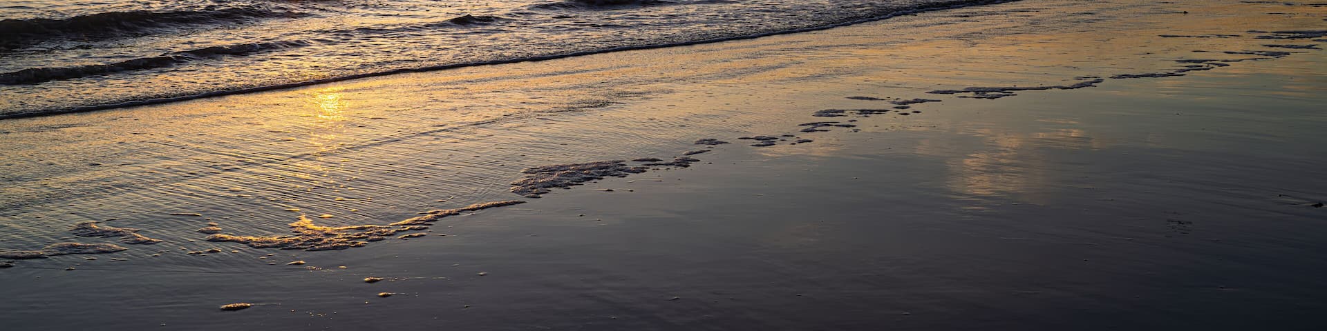 West St Leonards beach at sunset. The scene shows the beach at low ride, exposing the sand. Waves roll into the beach. The setting sun creates contrasting colours and deep shadows.