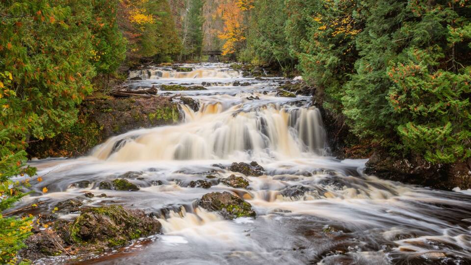 A multi-tiered waterfall surrounded by dense forest with autumn foliage.