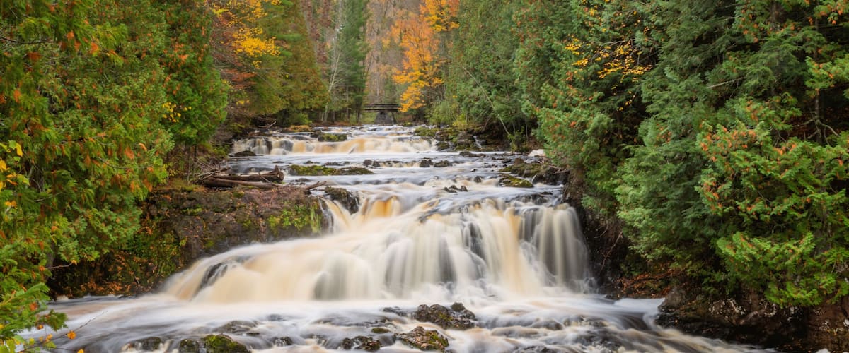 A multi-tiered waterfall surrounded by dense forest with autumn foliage.