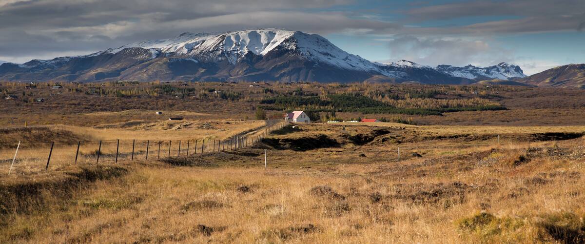 A snow-capped mountain range, a fenced field, and a small white building in a rural landscape.