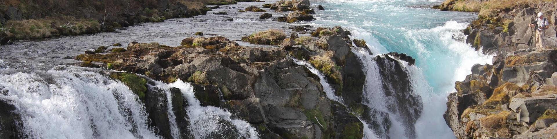 A waterfall cascading over rocks in a river surrounded by grassy hills.
