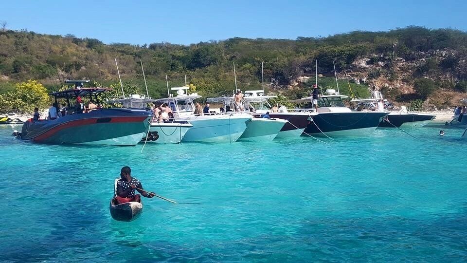 A row of boats docked in clear turquoise water with a person paddling a small boat in the foreground.