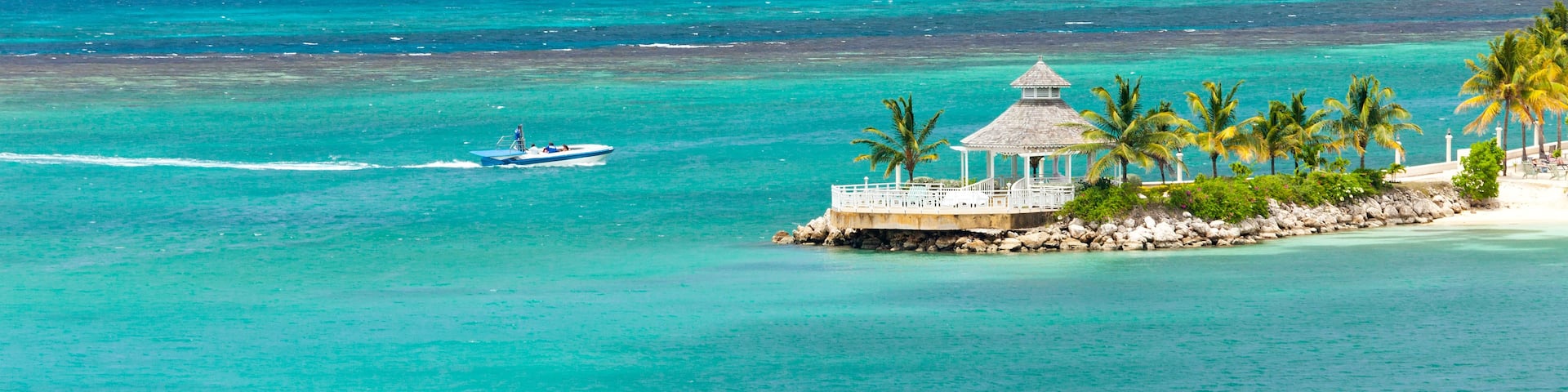 A clear blue sea with a parasail, a small boat, and a white gazebo with palm trees.