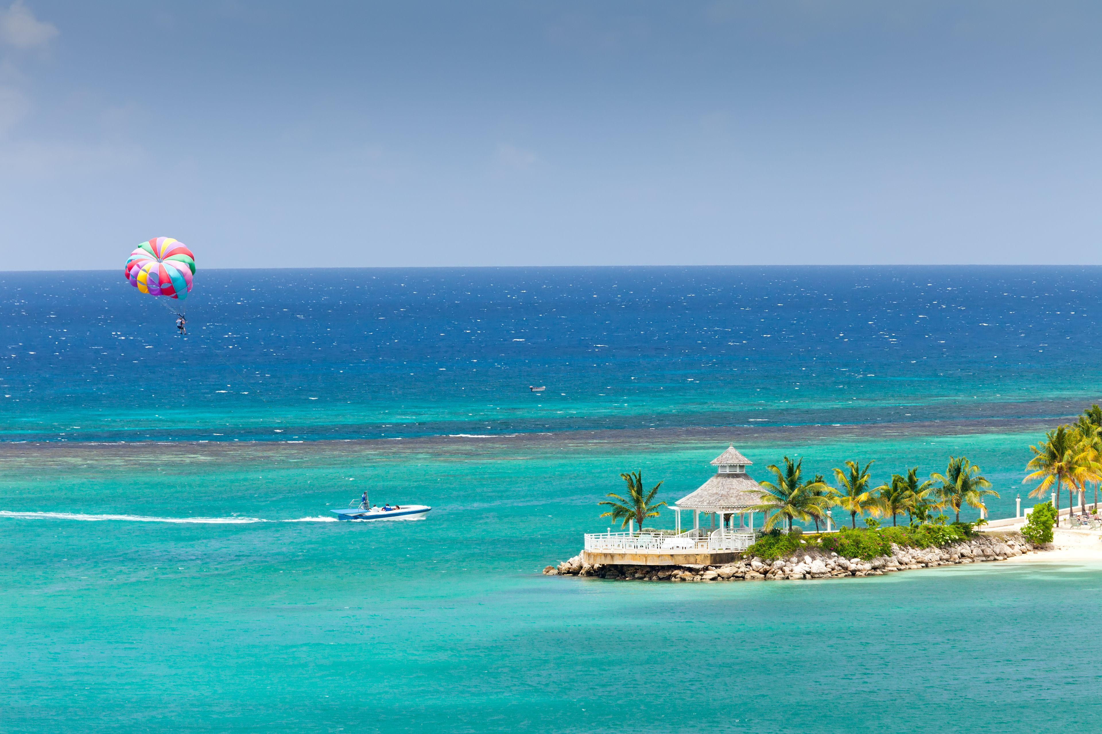 A clear blue sea with a parasail, a small boat, and a white gazebo with palm trees.
