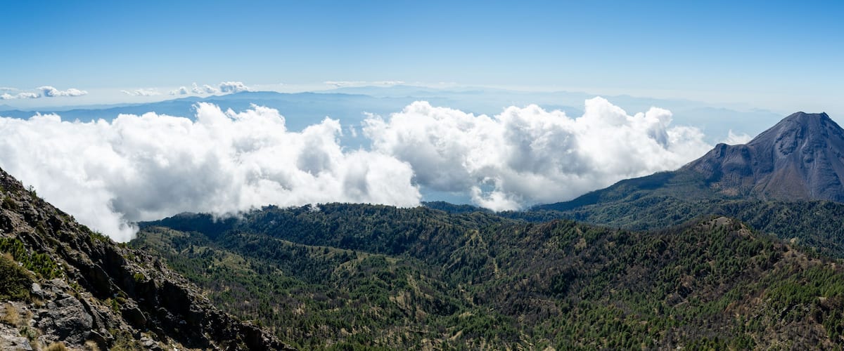 A mountain with a peak, surrounded by clouds and forested hills.