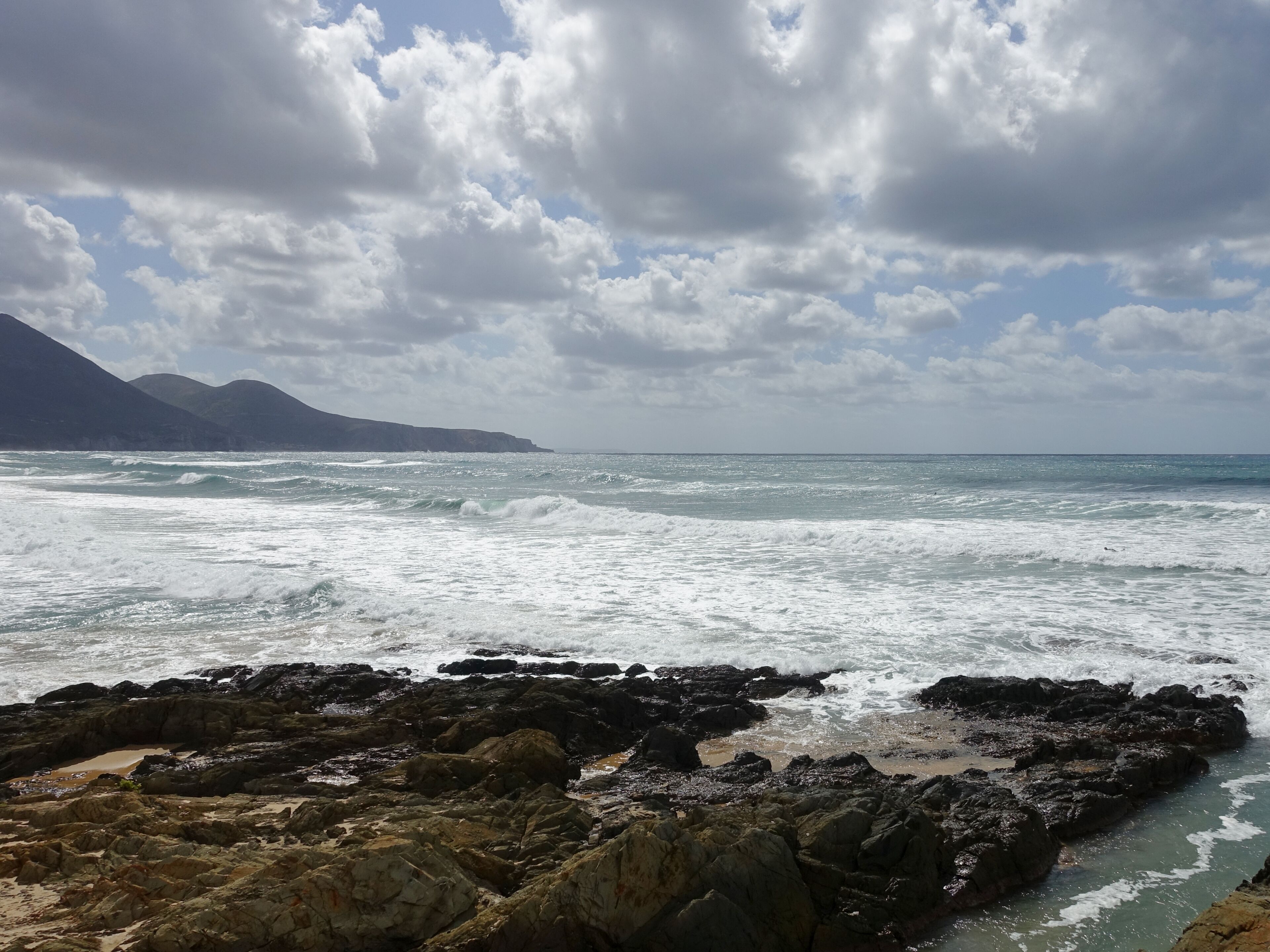 A rocky coastline with waves crashing against the shore and mountains in the background.