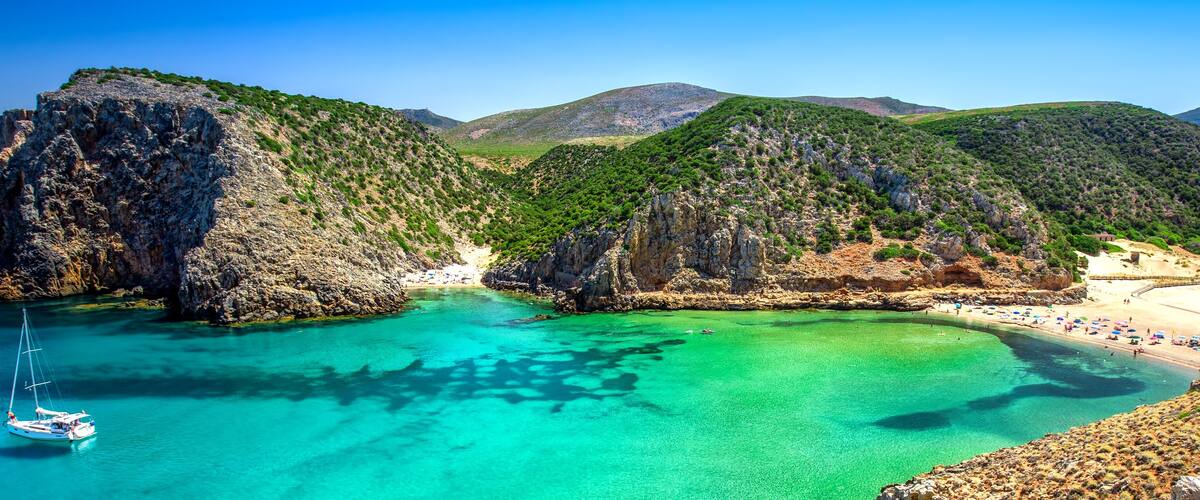 Paisatge costaner amb un mar turquesa transparent, penya-segats rocosos i una platja amb ombrel·les.