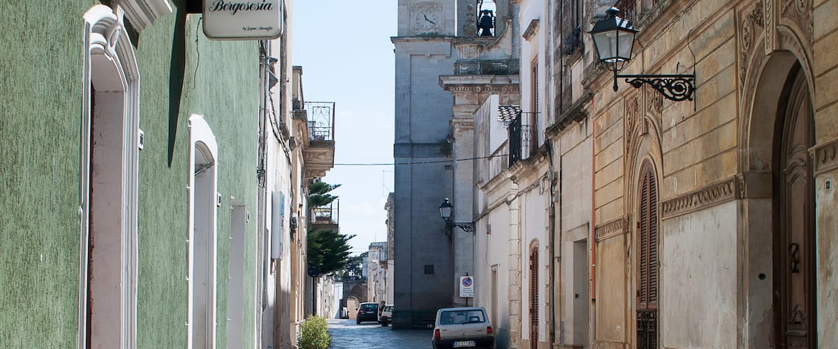 A narrow cobblestone street lined with historic buildings, a bell tower, and a sign for "La Bodega de la Reina."