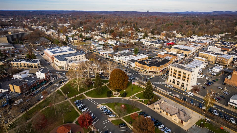 An aerial view of a town with a mix of commercial and residential buildings, a central roundabout, and surrounding green spaces.