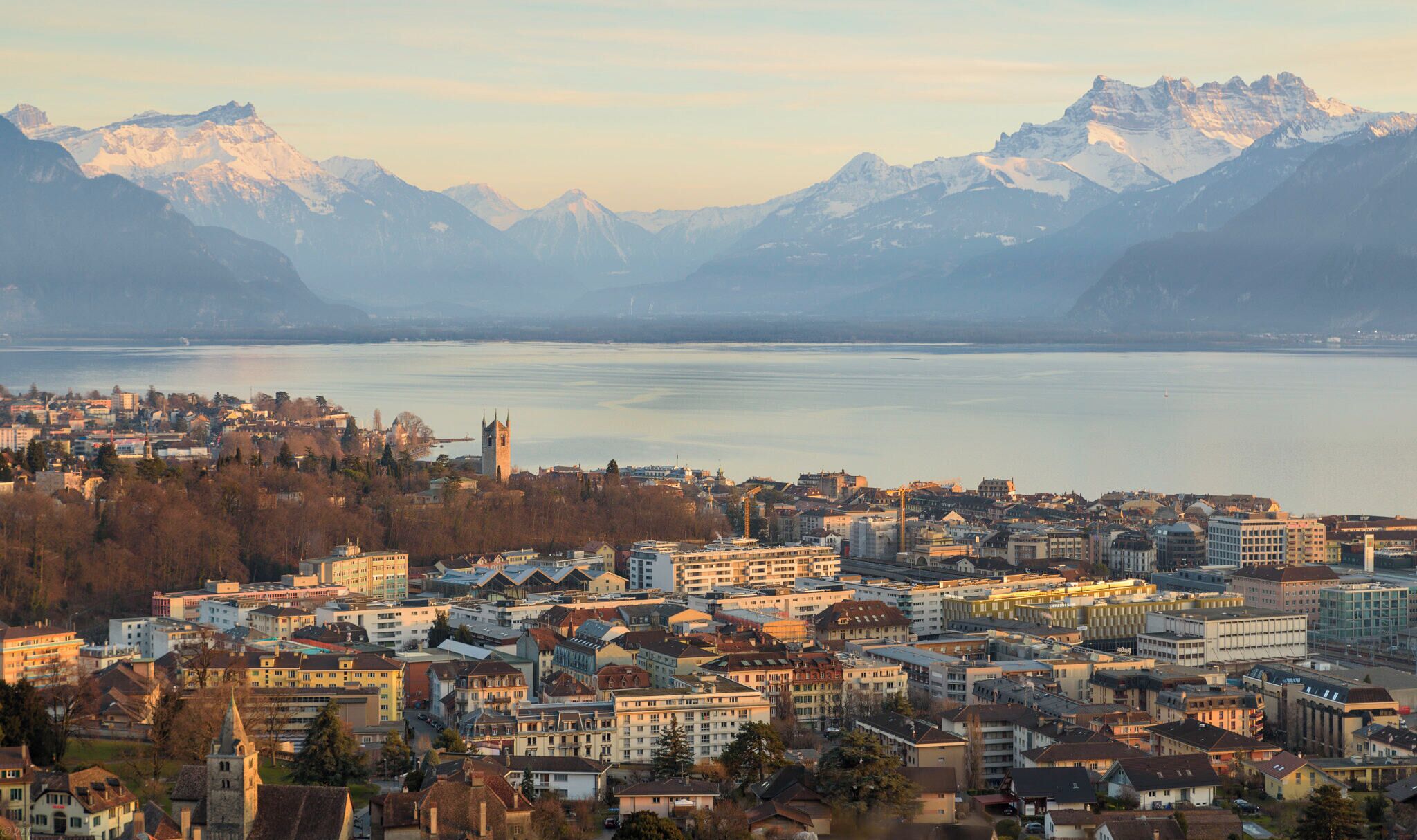 A cityscape with a prominent church tower, a lake, and snow-capped mountains in the background.