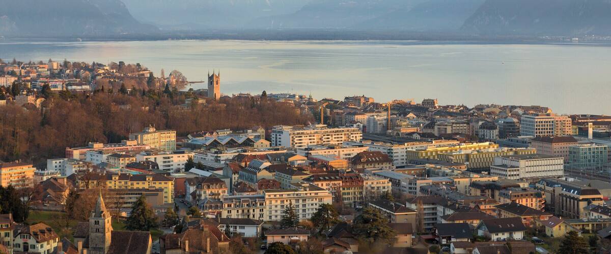 A cityscape with a prominent church tower, a lake, and snow-capped mountains in the background.