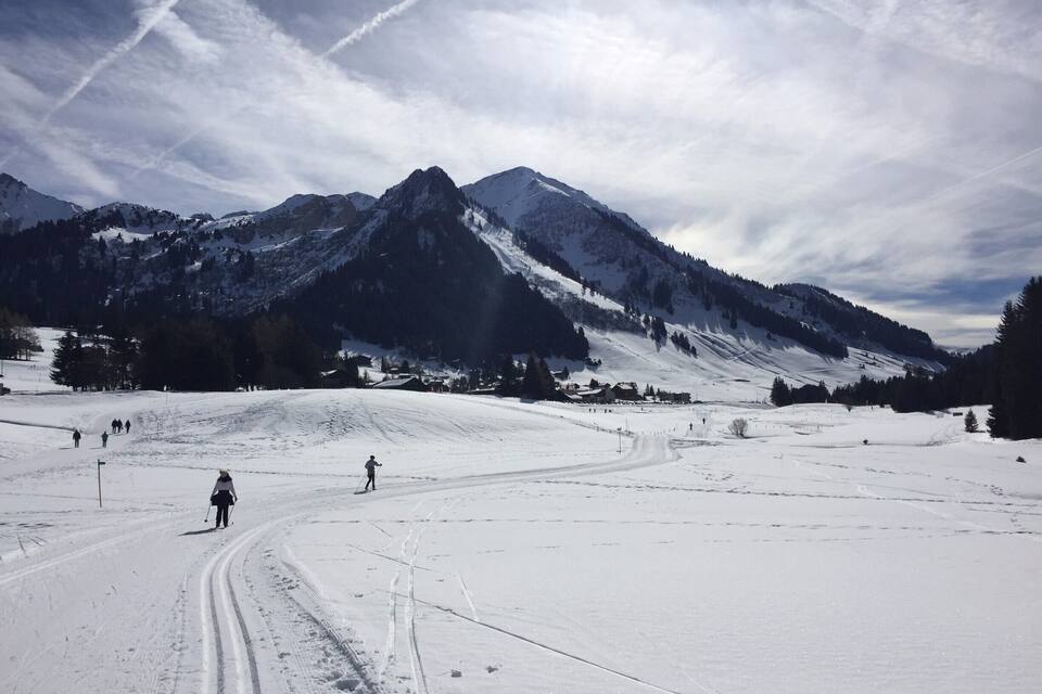 Une piste de ski enneigée avec des skieurs, une chaîne de montagnes et un ciel partiellement nuageux.