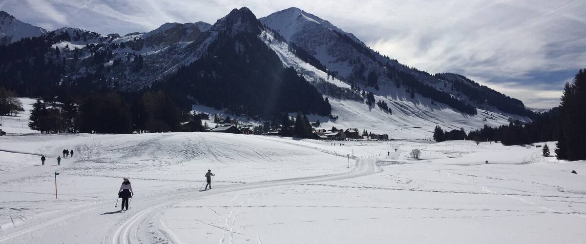 A snow-covered ski slope with skiers, a mountain range, and a partly cloudy sky.