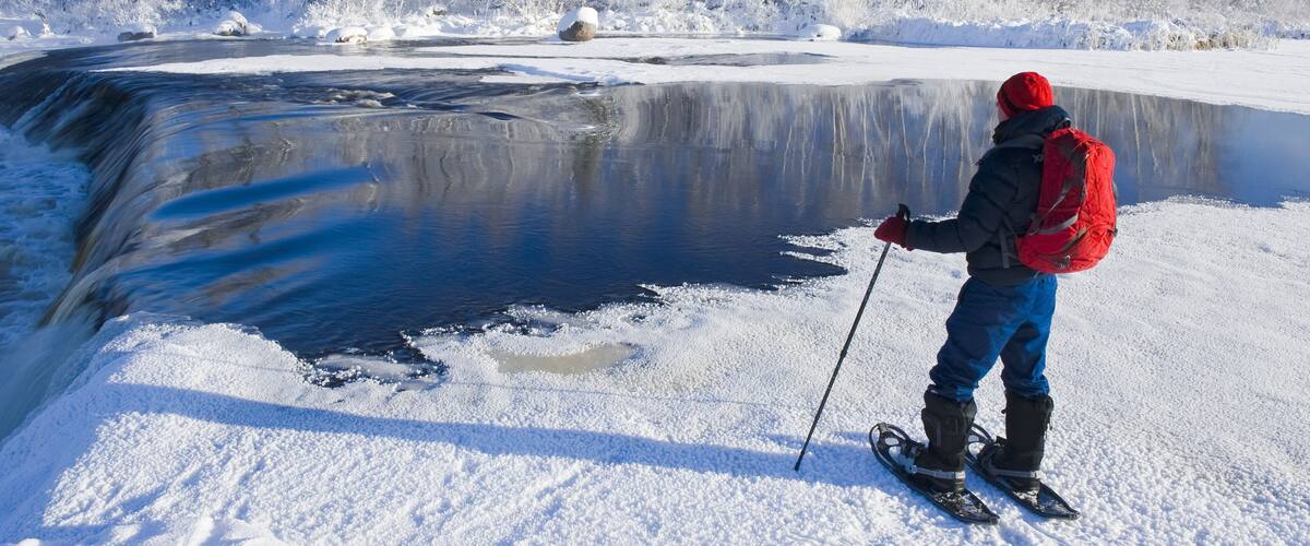 A person on skis near a partially frozen river with snow-covered trees in the background.