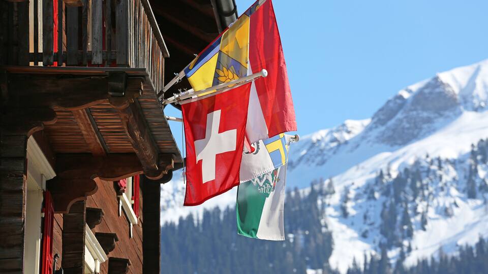 A wooden building with red shutters and flags, including the Swiss flag, against a snowy mountain backdrop.