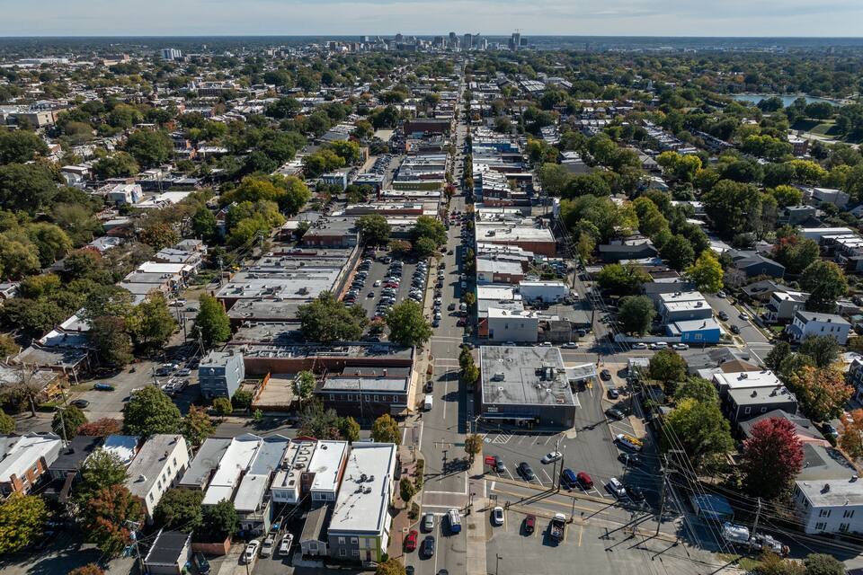 An aerial view of a densely populated urban area with a mix of residential and commercial buildings, a main road, and a distant city skyline.