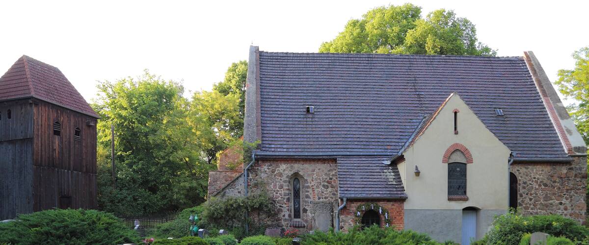 A church with a stone facade and a tiled roof, surrounded by lush greenery and a cemetery.