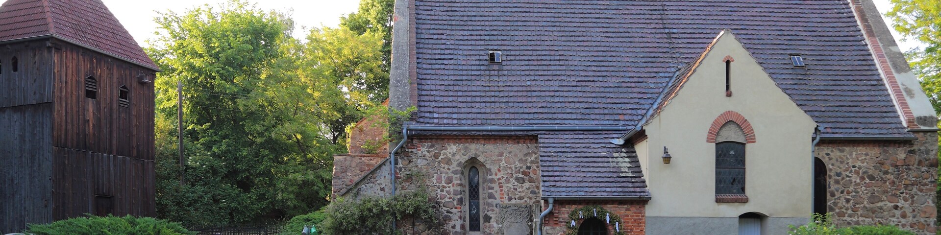 A church with a stone facade and a tiled roof, surrounded by lush greenery and a cemetery.