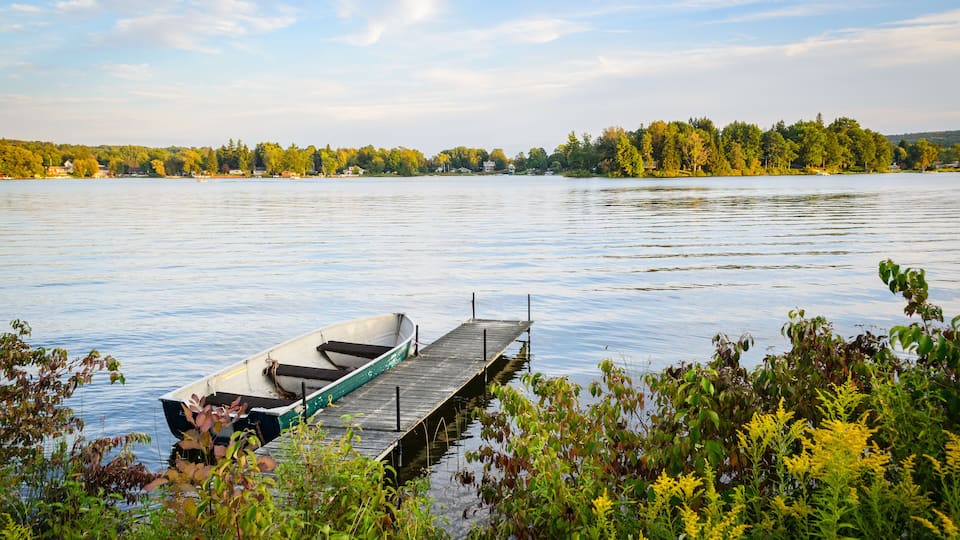A calm lake with a small boat docked at a wooden pier, surrounded by lush greenery and trees in the background.