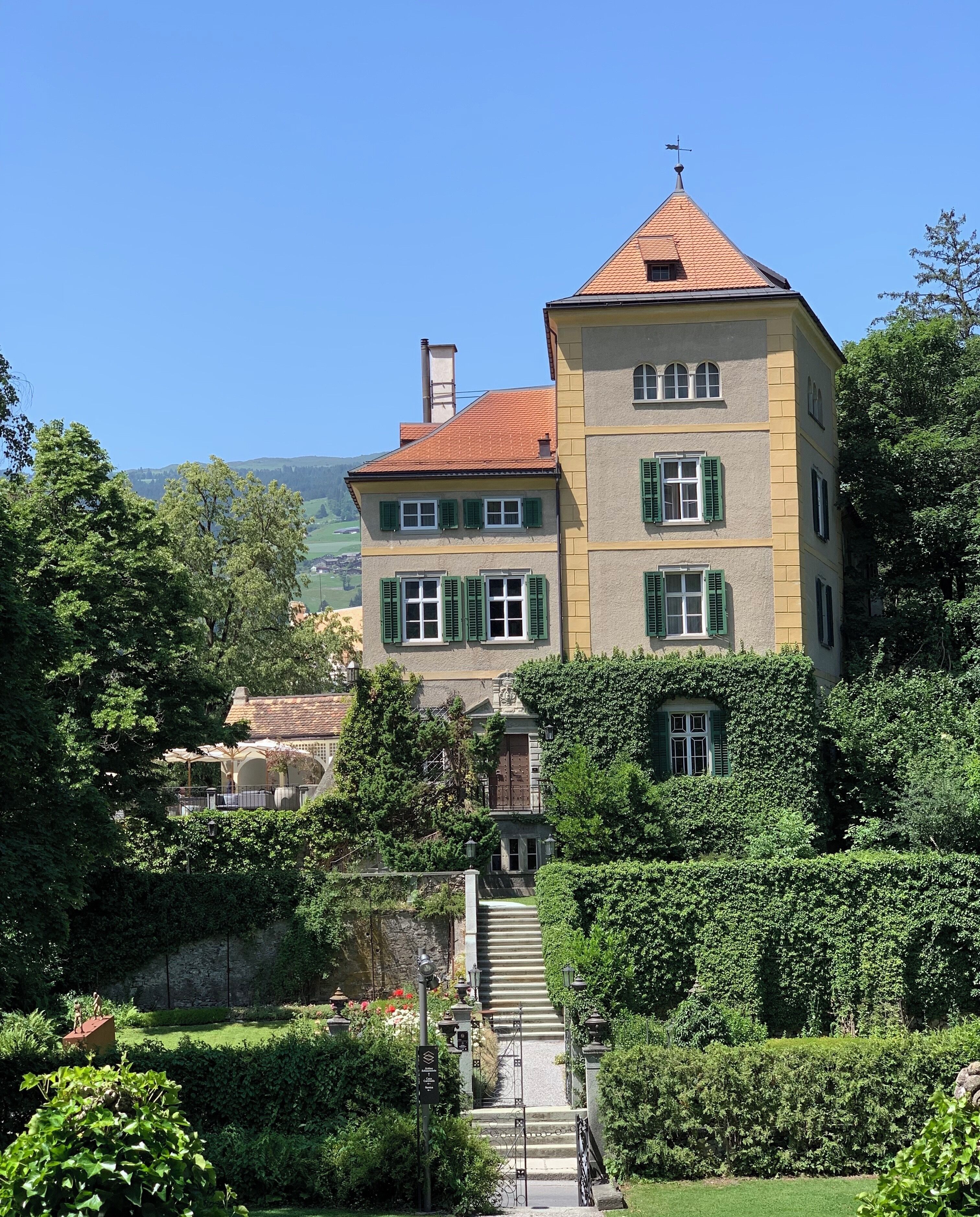 A large, multi-story building with a red-tiled roof, surrounded by lush greenery and a well-maintained garden.