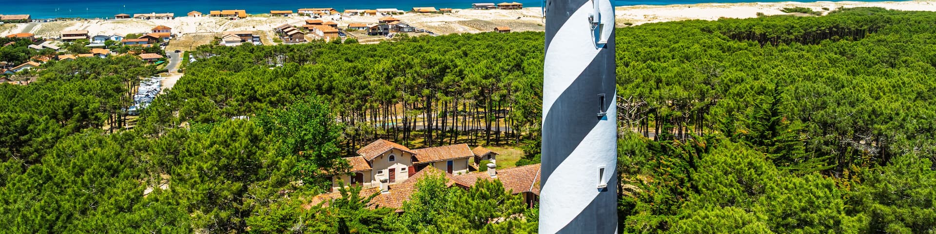 A lighthouse surrounded by dense pine forests with a beach and coastal town in the background.