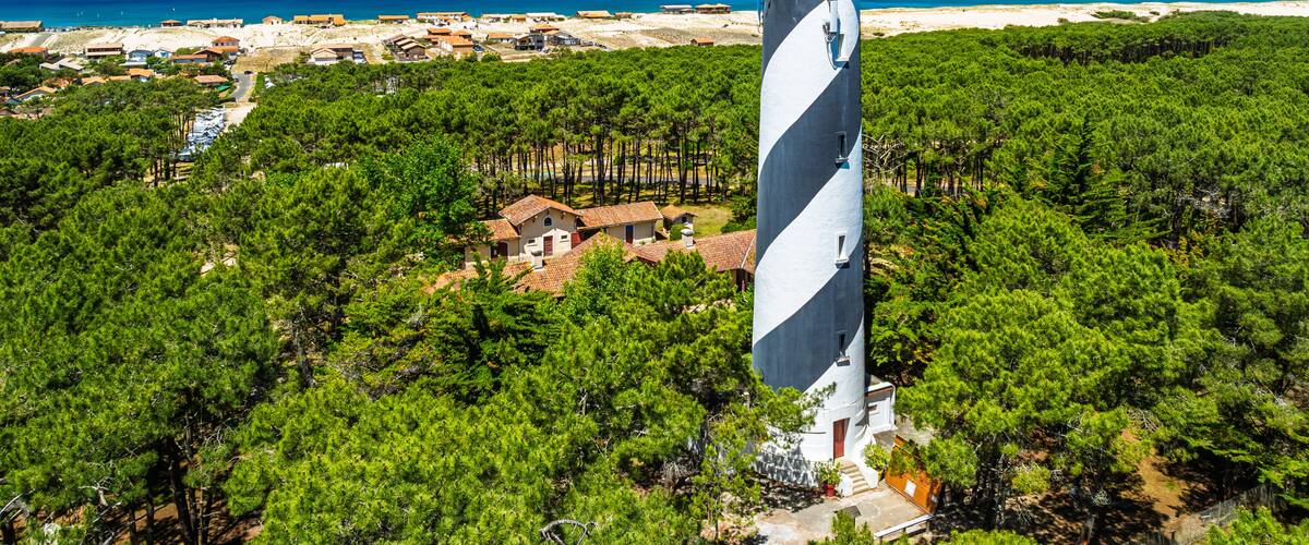 A lighthouse surrounded by dense pine forests with a beach and coastal town in the background.