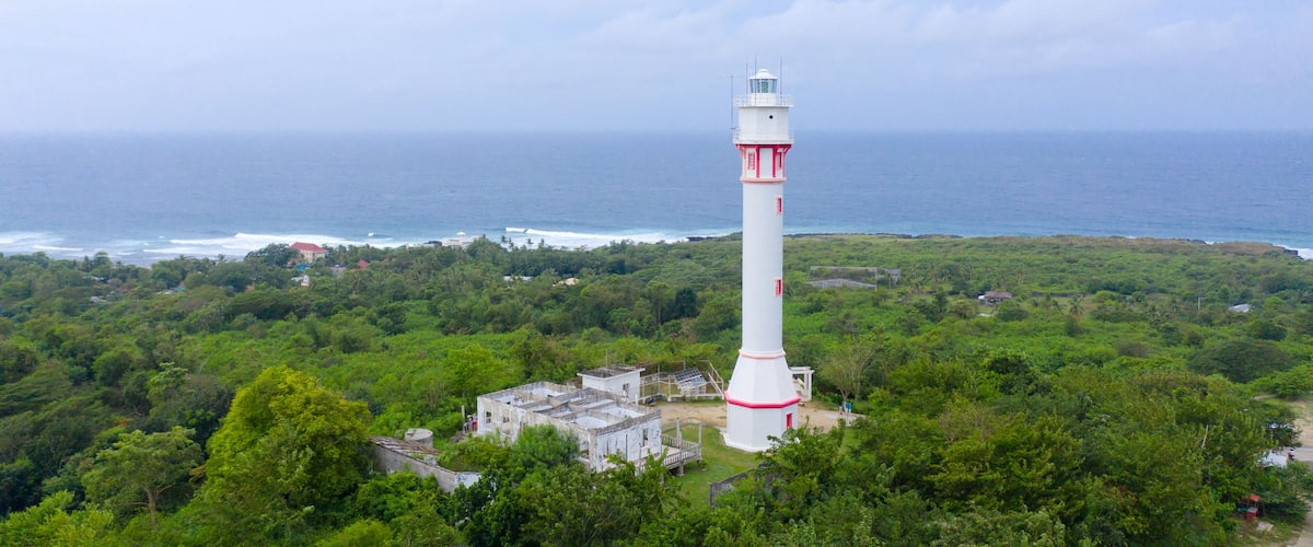 A white lighthouse with red stripes, surrounded by lush greenery and overlooking the ocean.
