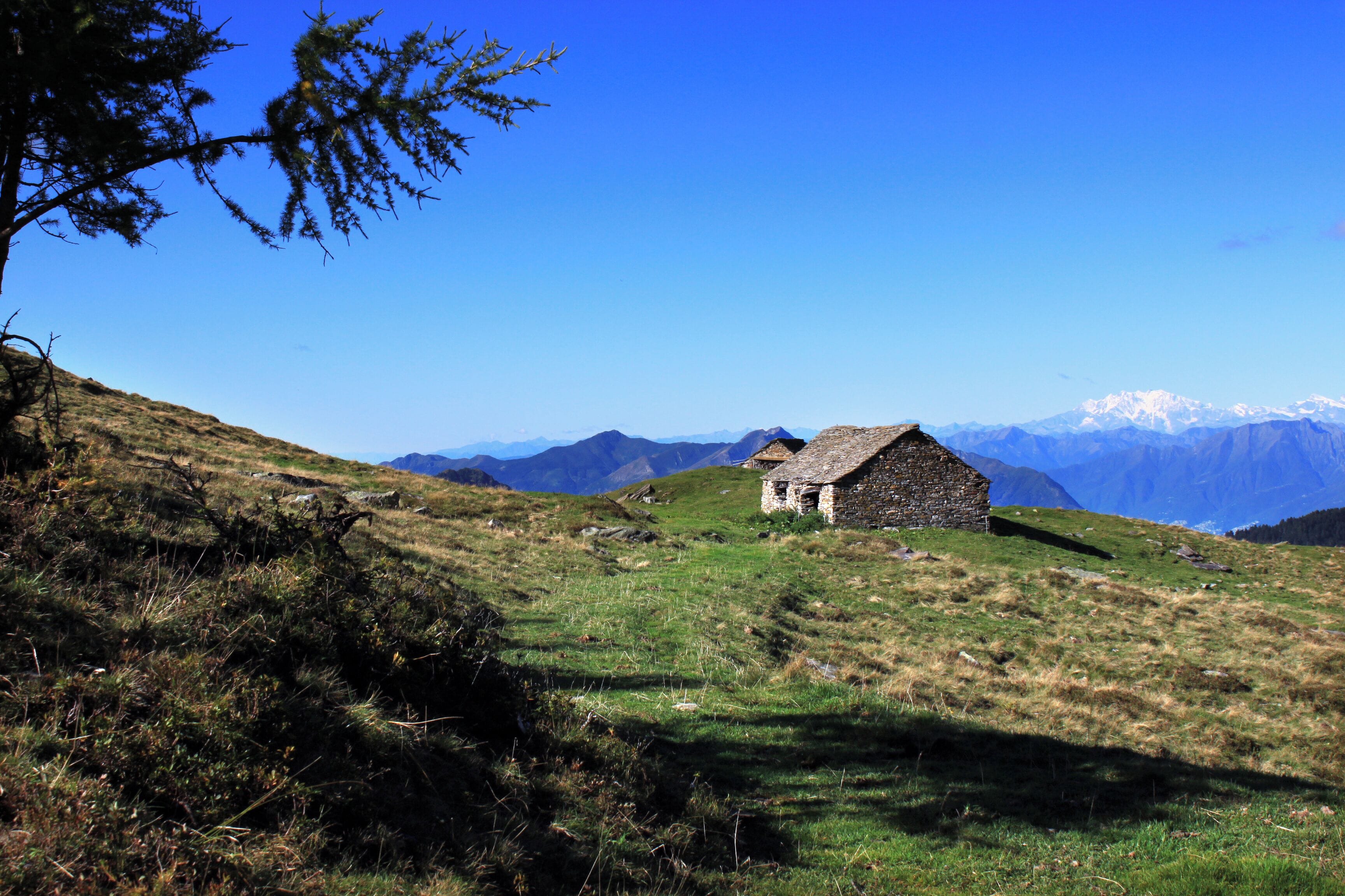A stone house on a grassy hillside with a clear blue sky and snow-capped mountains in the background.